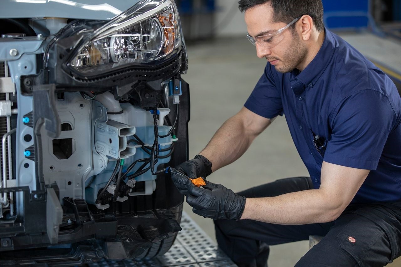 Mechanic in safety glasses and gloves working on the front wiring of a car with its bumper removed.