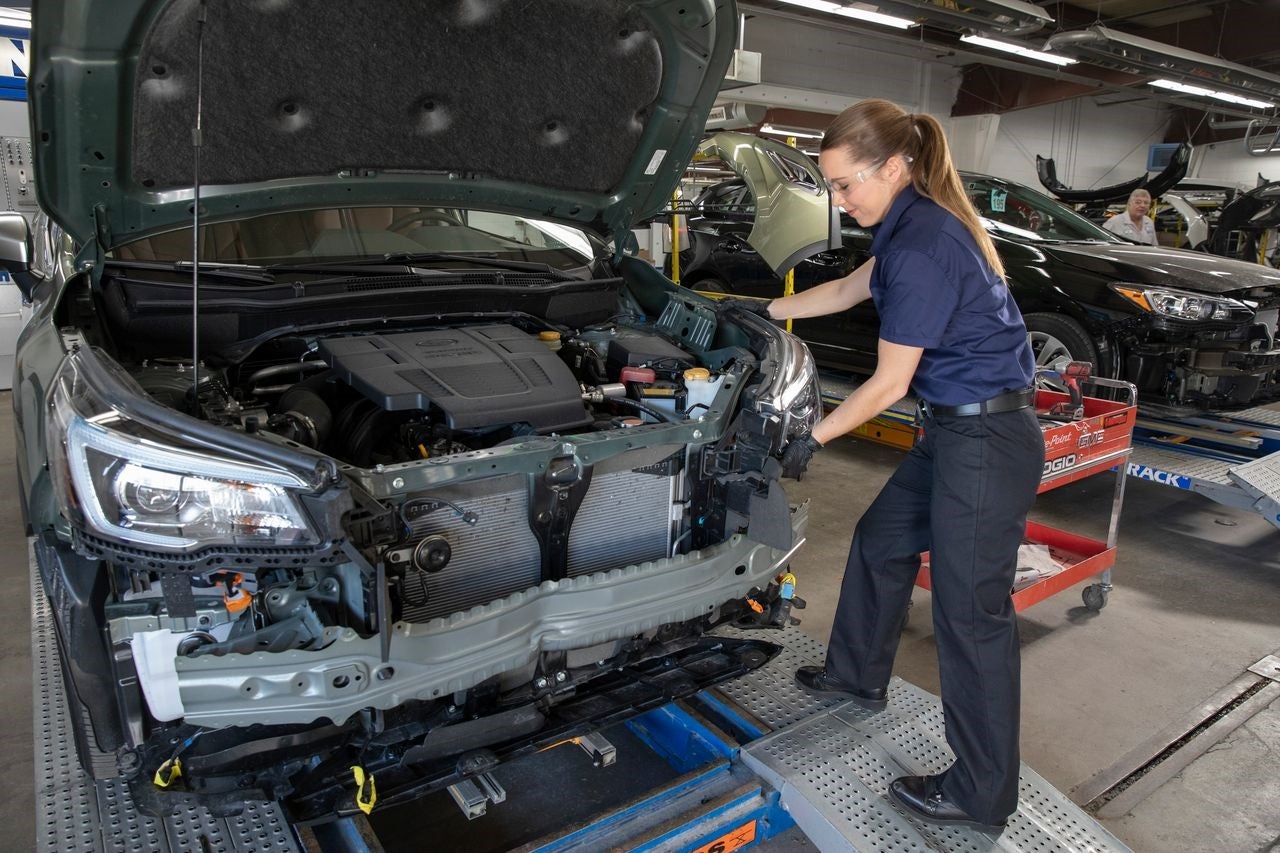 A female auto technician in safety glasses working on the front of a partially disassembled green car in a garage.