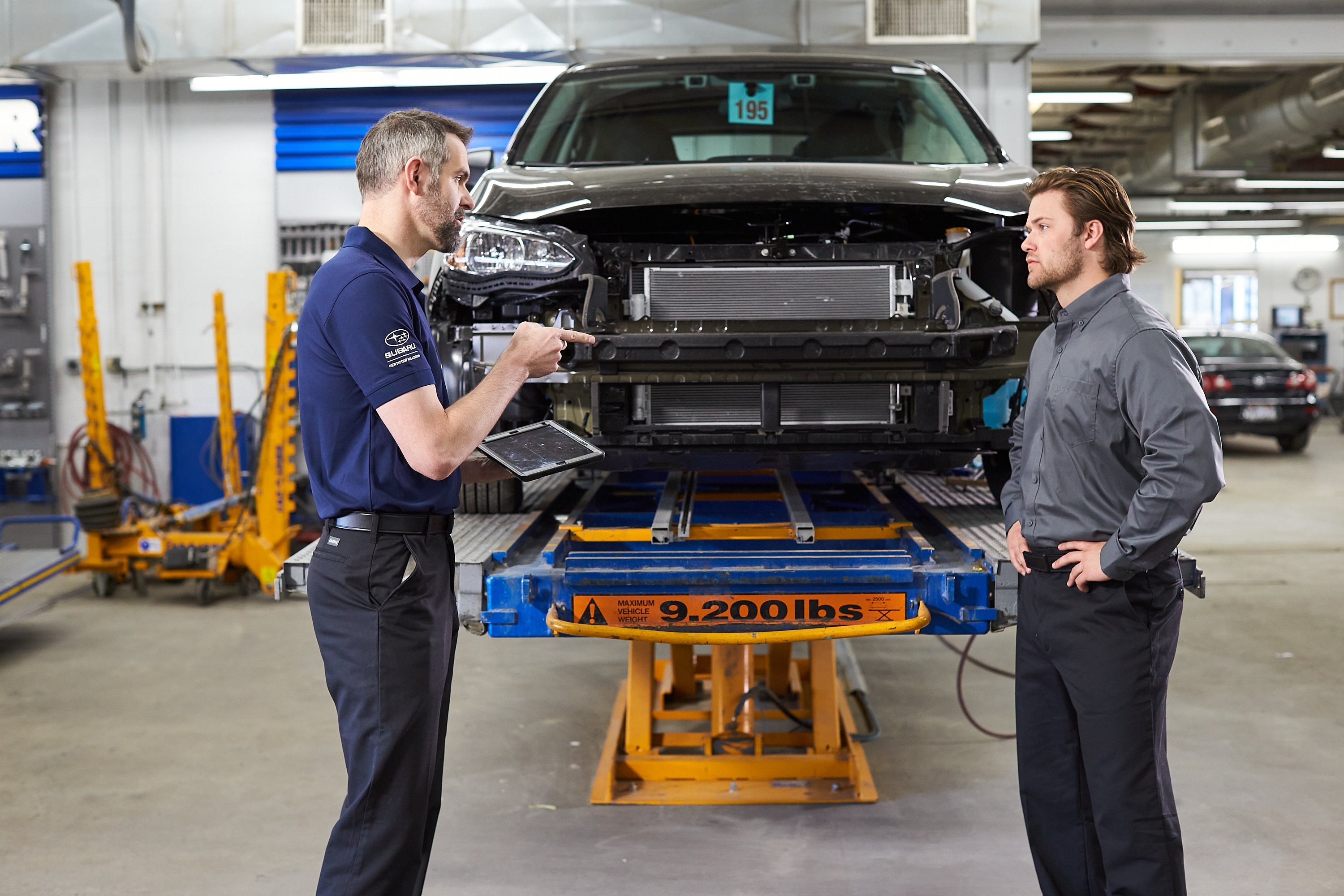 Two men in a garage discussing a car with its front bumper removed, sitting on a hydraulic lift.