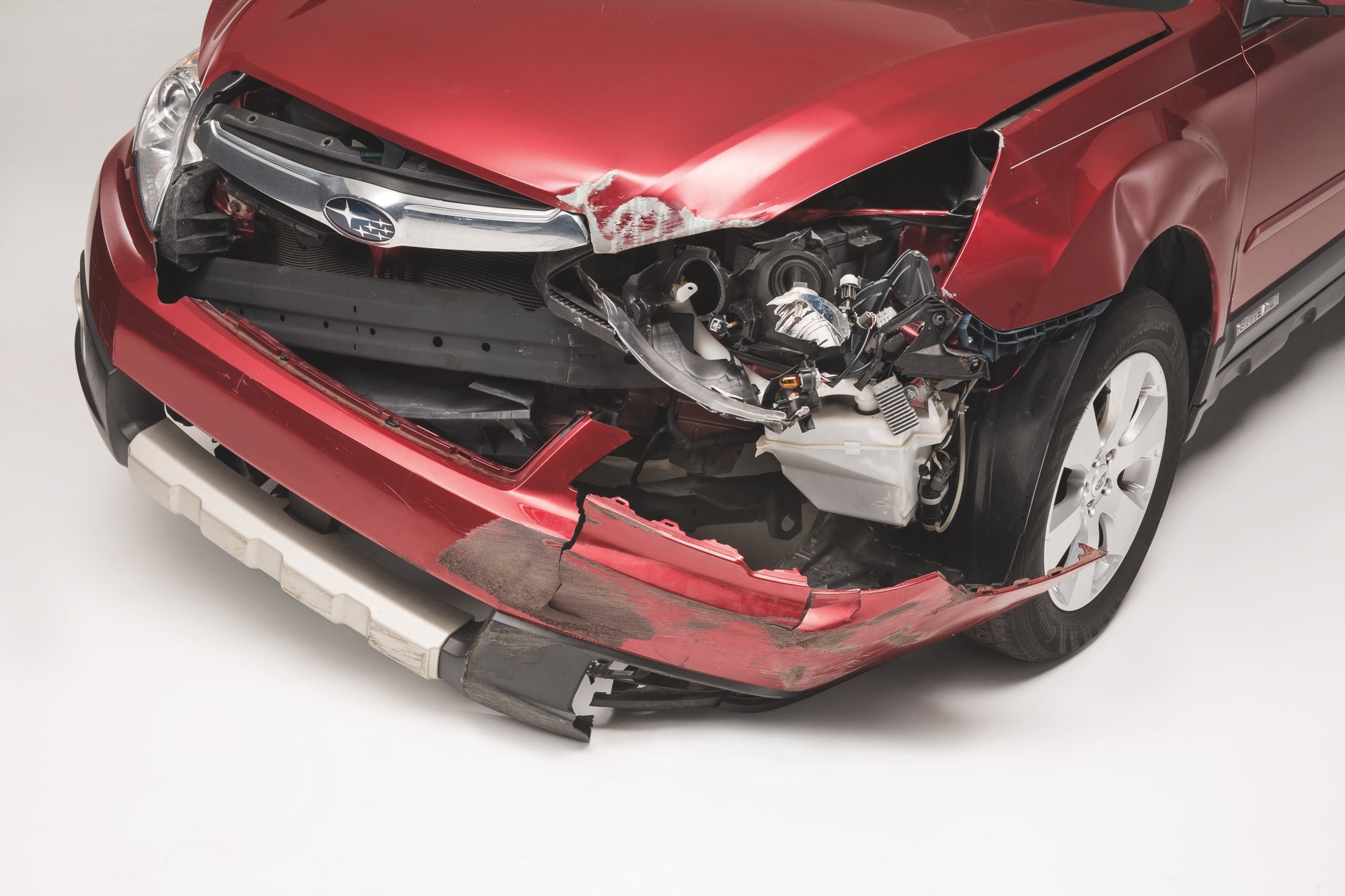 Front view of a damaged red Subaru car with the bumper torn off and the passenger-side headlight broken.
