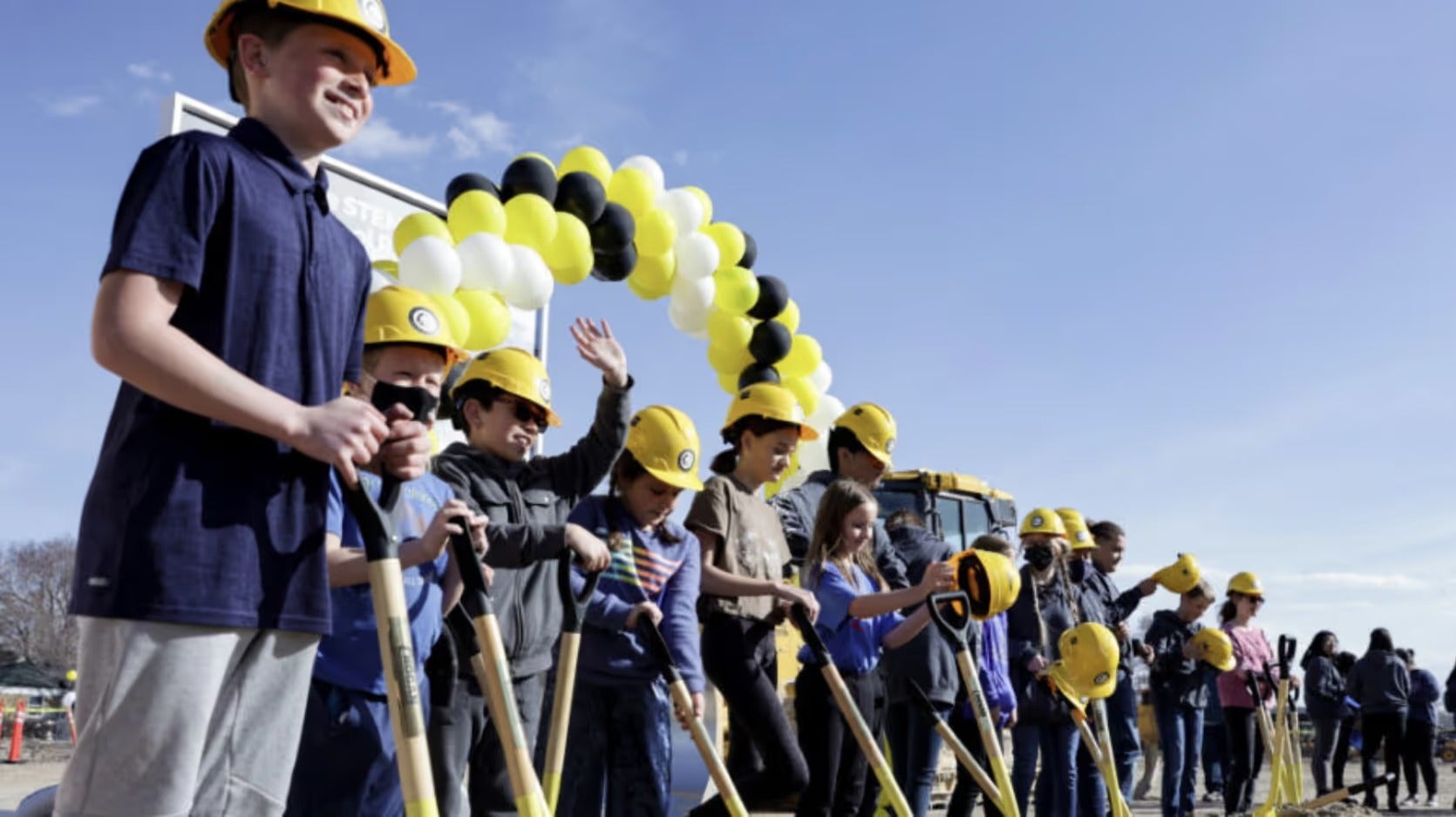 Children in yellow hard hats and dark shirts hold shovels at a groundbreaking ceremony with a balloon arch.